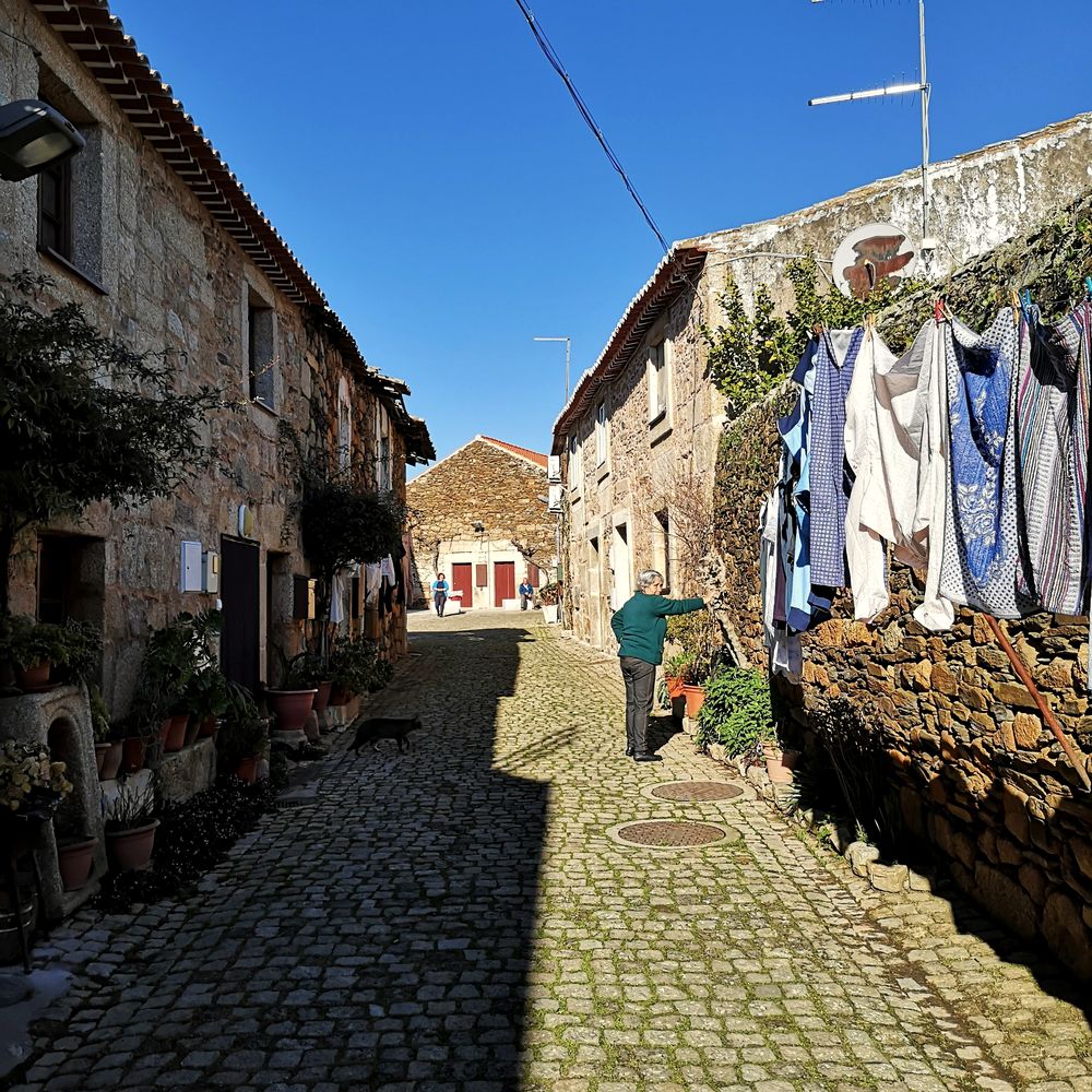Calle empedrada de un pueblo de piedra con ropa tendida y una vecina regando las plantas